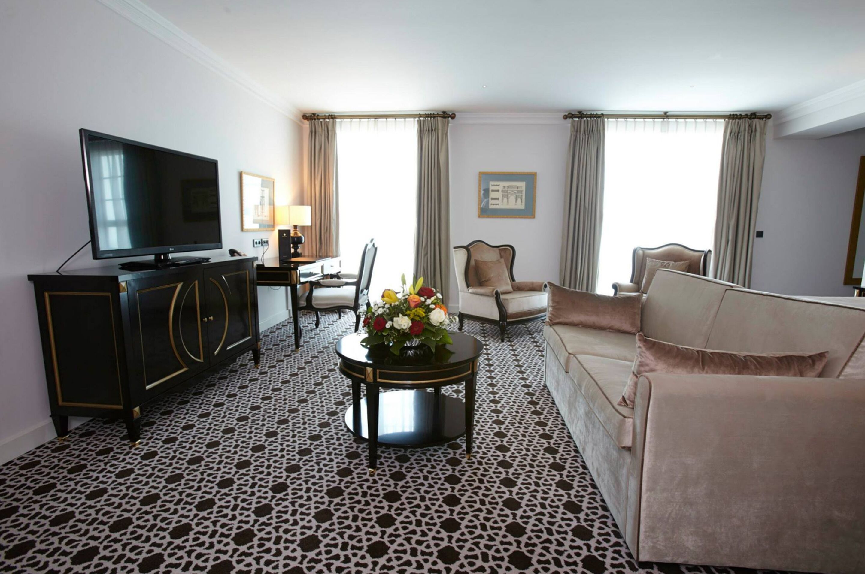 Une chambre d’hôtel dotée d’un téléviseur à écran plat, d’un tapis à motifs noir et blanc, d’un canapé beige, d’une table basse ronde agrémentée d’un arrangement floral et d’un bureau avec une lampe.