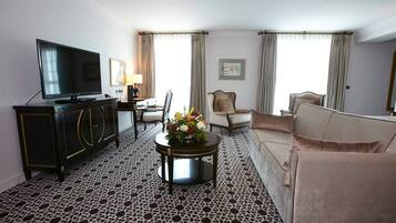 Une chambre d’hôtel dotée d’un téléviseur à écran plat, d’un tapis à motifs noir et blanc, d’un canapé beige, d’une table basse ronde agrémentée d’un arrangement floral et d’un bureau avec une lampe.