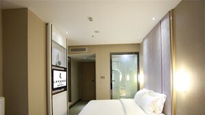 Habitación de hotel con cama, mesita de noche y un espejo. Se ve una puerta que da a un baño y un pasillo.
