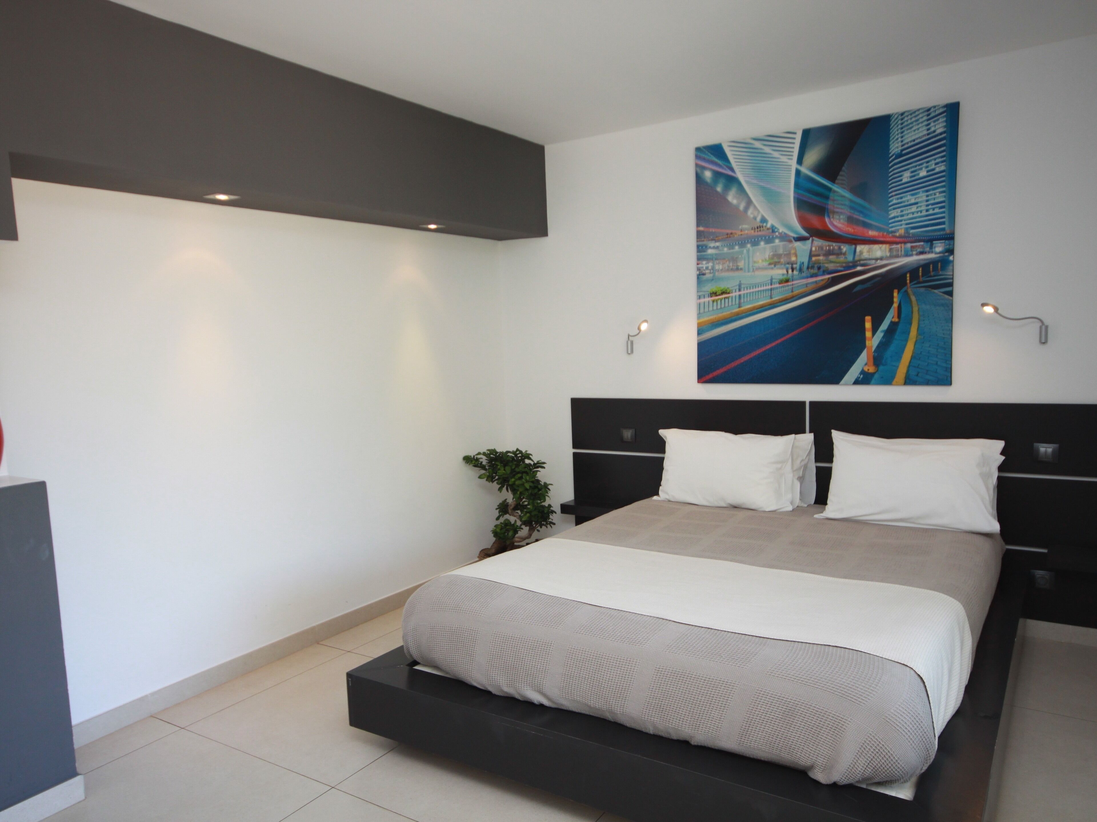 A modern bedroom with a large bed, a cityscape painting, and a small plant.