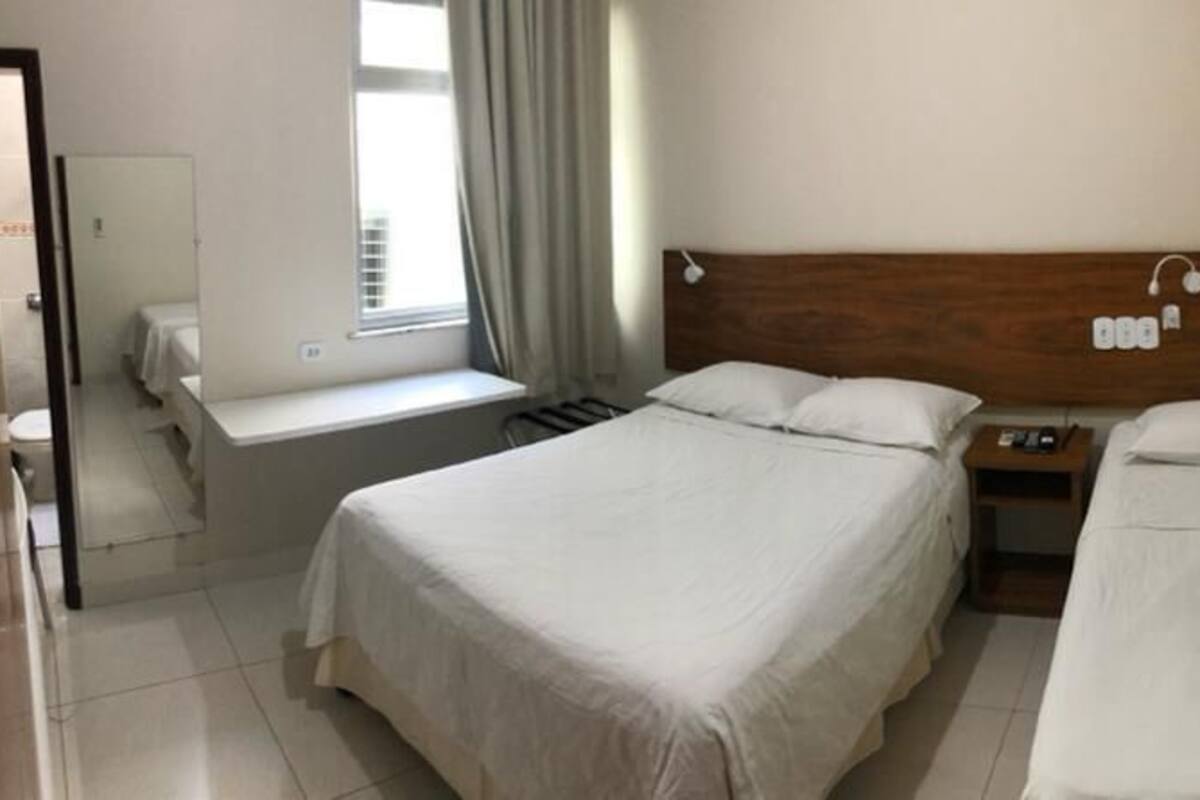 Une chambre d’hôtel avec deux lits simples, une tête de lit en bois, une table de chevet avec une lampe et une fenêtre avec des rideaux.