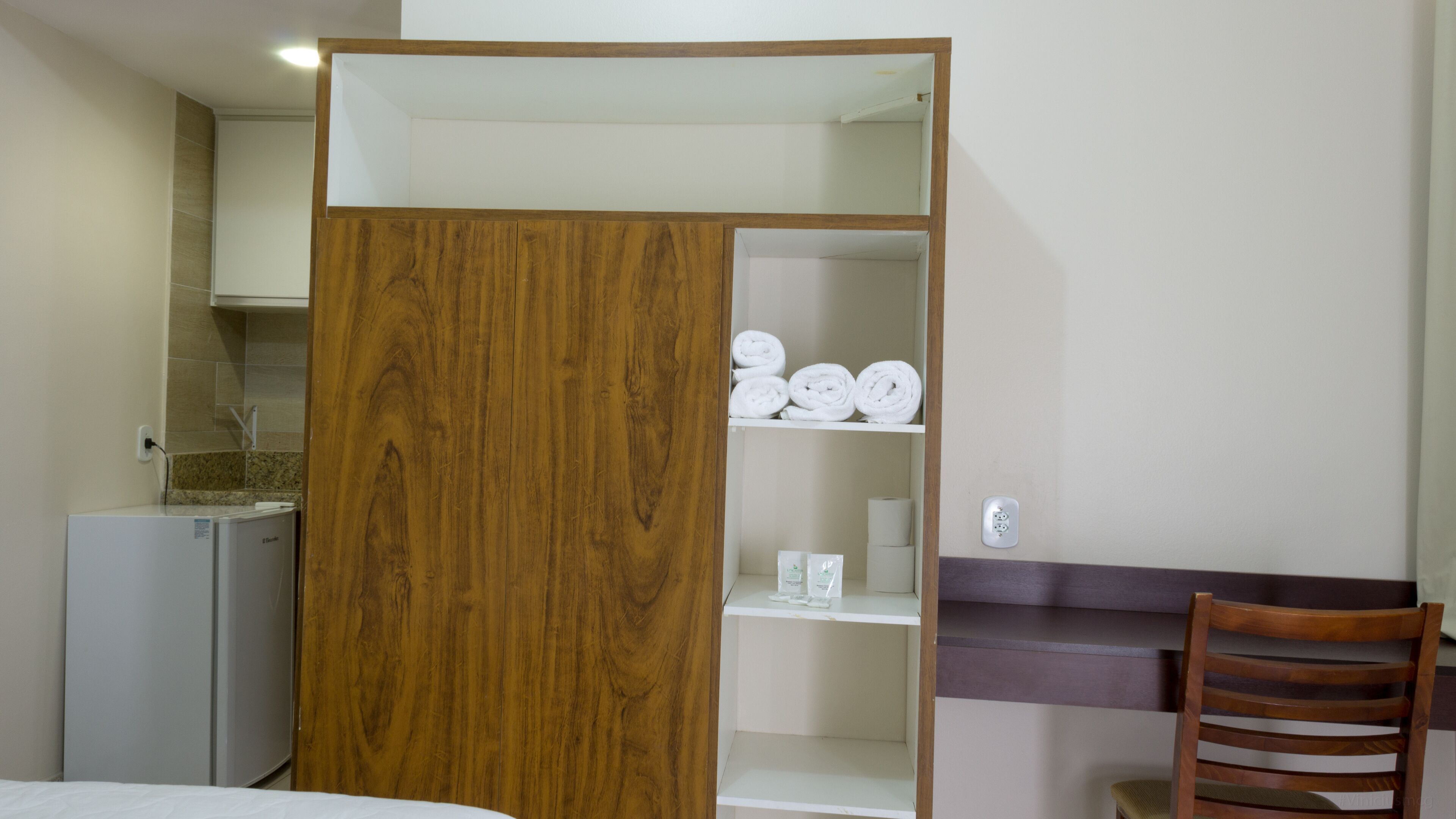 A hotel room with a wooden wardrobe, a bed, a desk with a chair, and a kitchenette area.