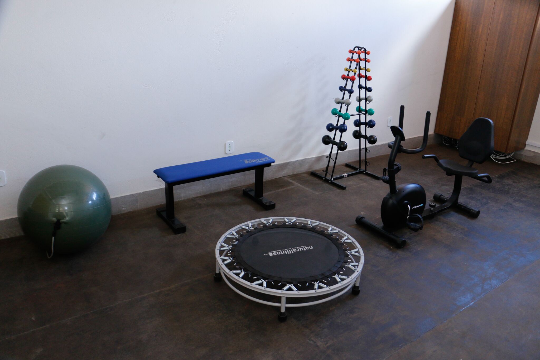Un gimnasio en casa con un banco azul, una pelota grande de ejercicios, un trampolín, un soporte para mancuernas y una bicicleta estática.