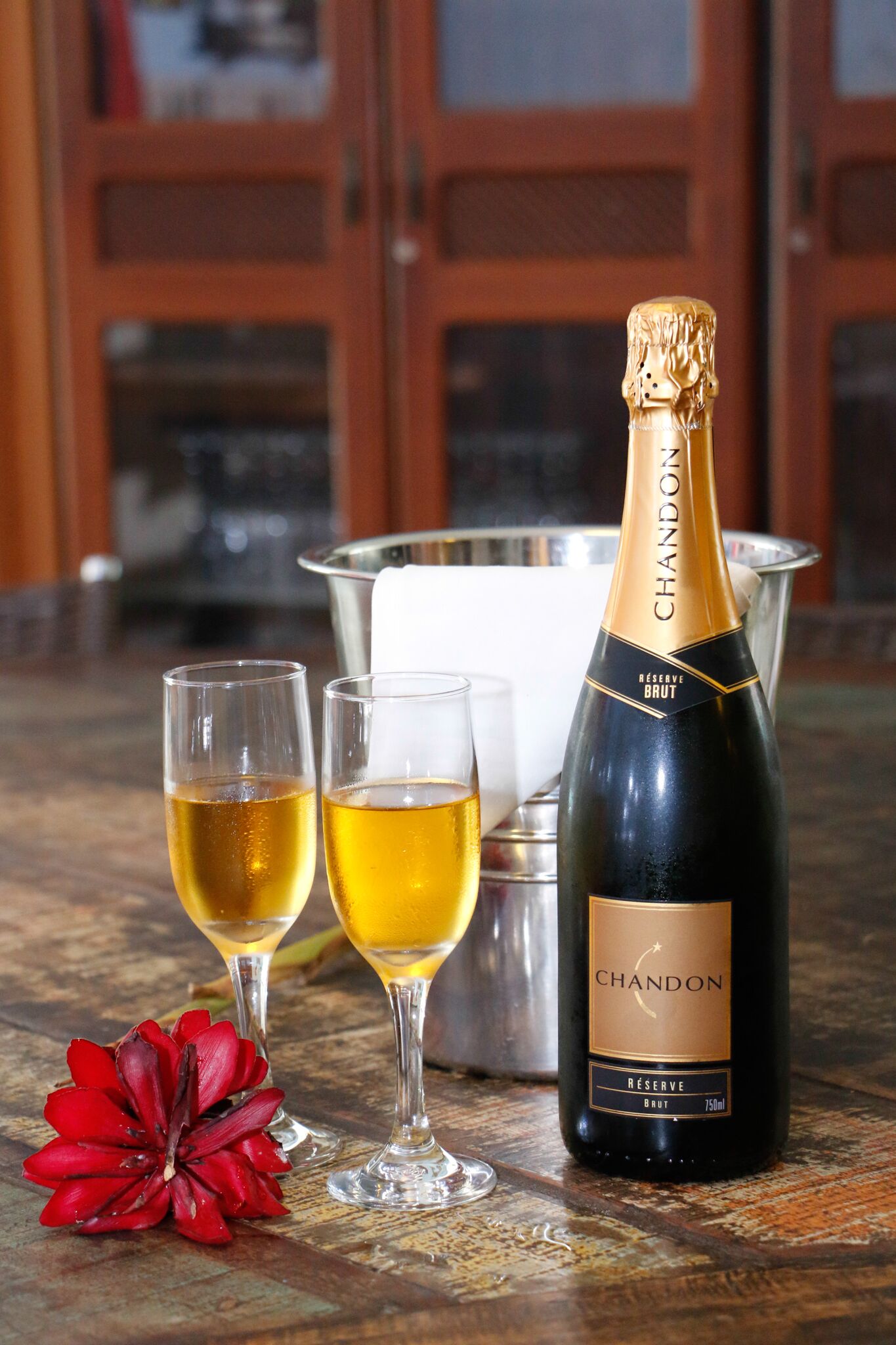 Una botella de champagne Chandon Reserve Brut, dos copas de champagne y una flor roja sobre una mesa de madera.