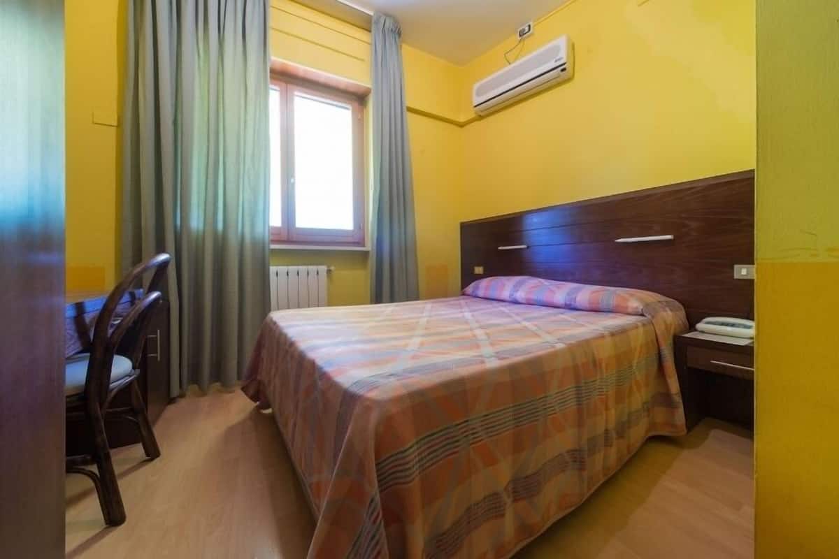 Una habitación con cama, escritorio y silla. Se ve una ventana con cortinas, un equipo de aire acondicionado en la pared y un radiador debajo de la ventana.