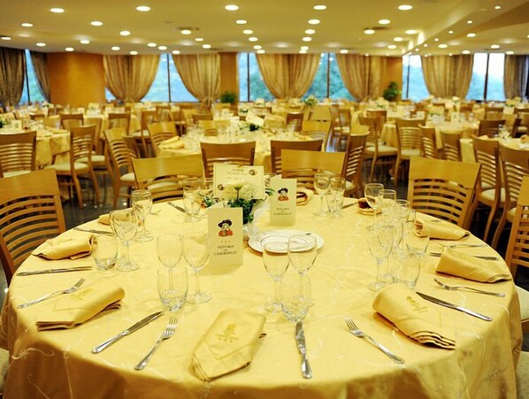 Uma sala de banquetes com mesas redondas dispostas para refeições, cada uma com um cardápio, copos de vinho e talheres.