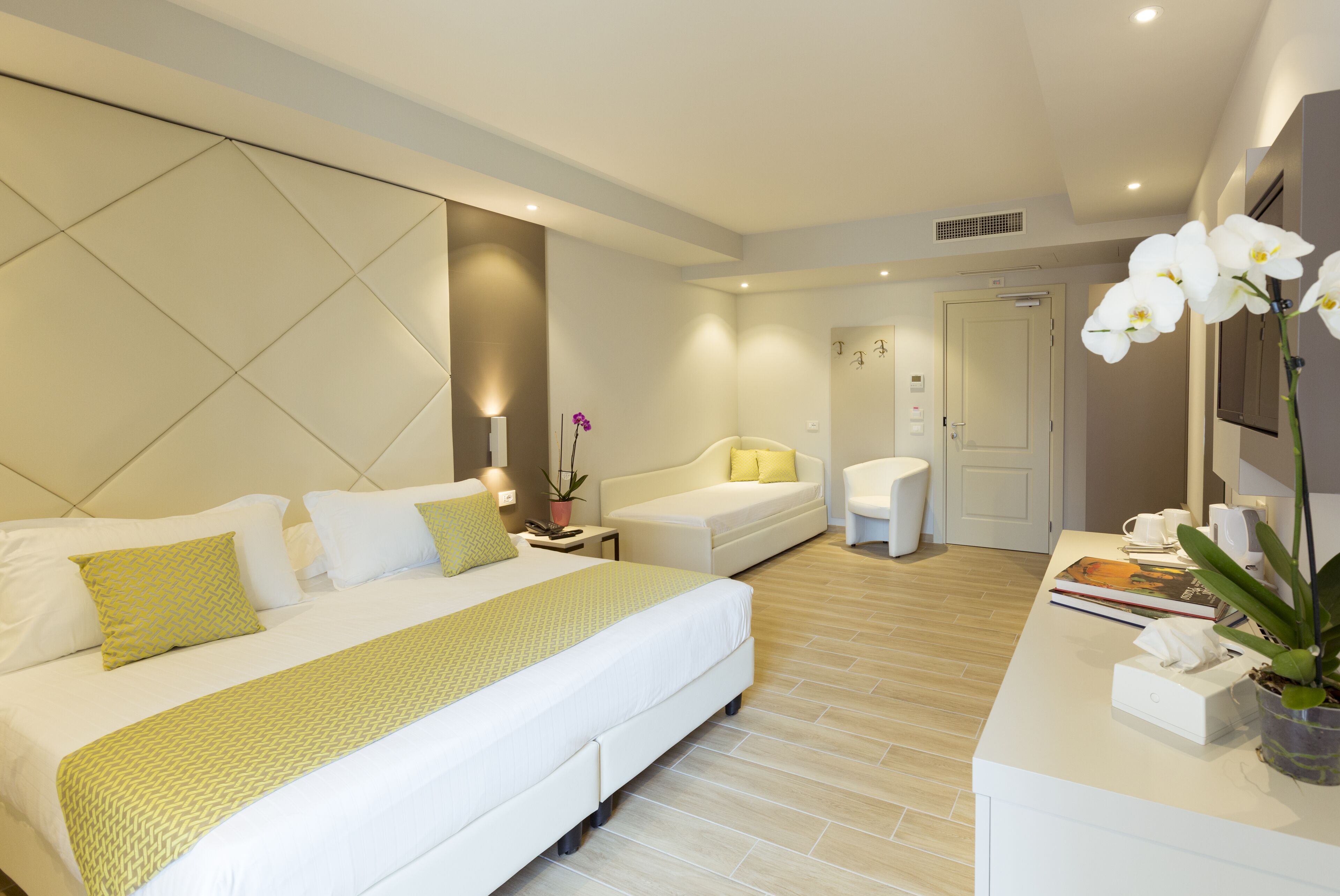 Ein modernes Hotelzimmer mit einem großen Bett, einem Nachttisch mit Lampe, einer kleinen Couch und einem Schreibtisch mit Computer und Topfpflanze.