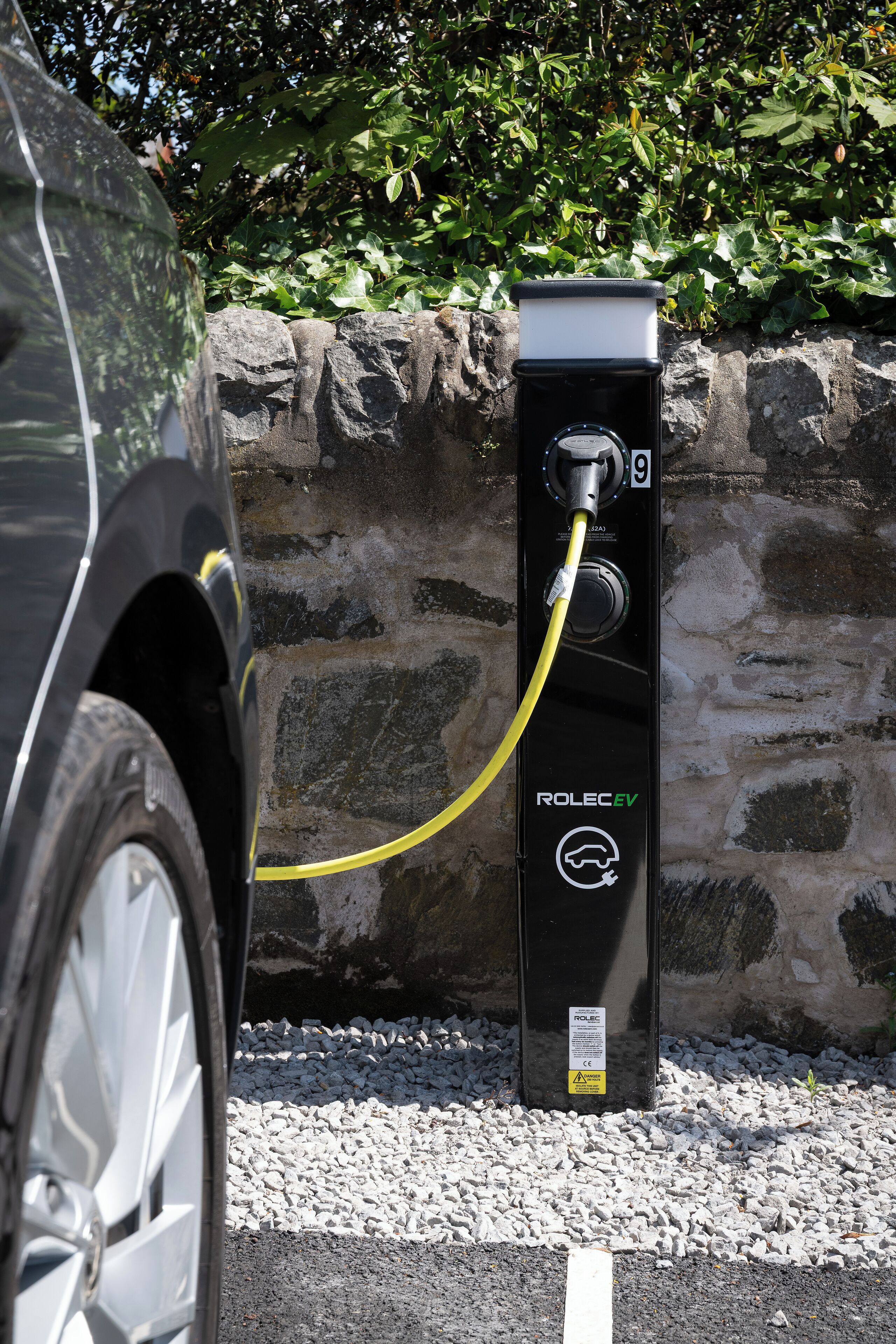 En sort elbil-ladestation med teksten 'ROLEC EV', med et gult ladekabel tilsluttet en bil.