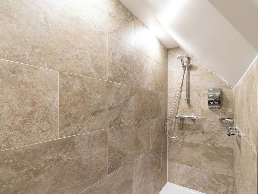 Un baño moderno con azulejos color beige, una bañera blanca y una ducha con cabezal de ducha manual.
