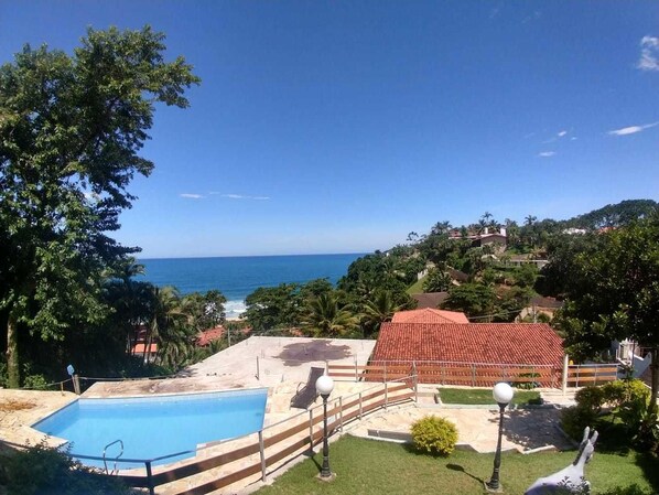 Área da piscina com vista clara para o oceano, cercada por vegetação e um edifício com telhado vermelho.