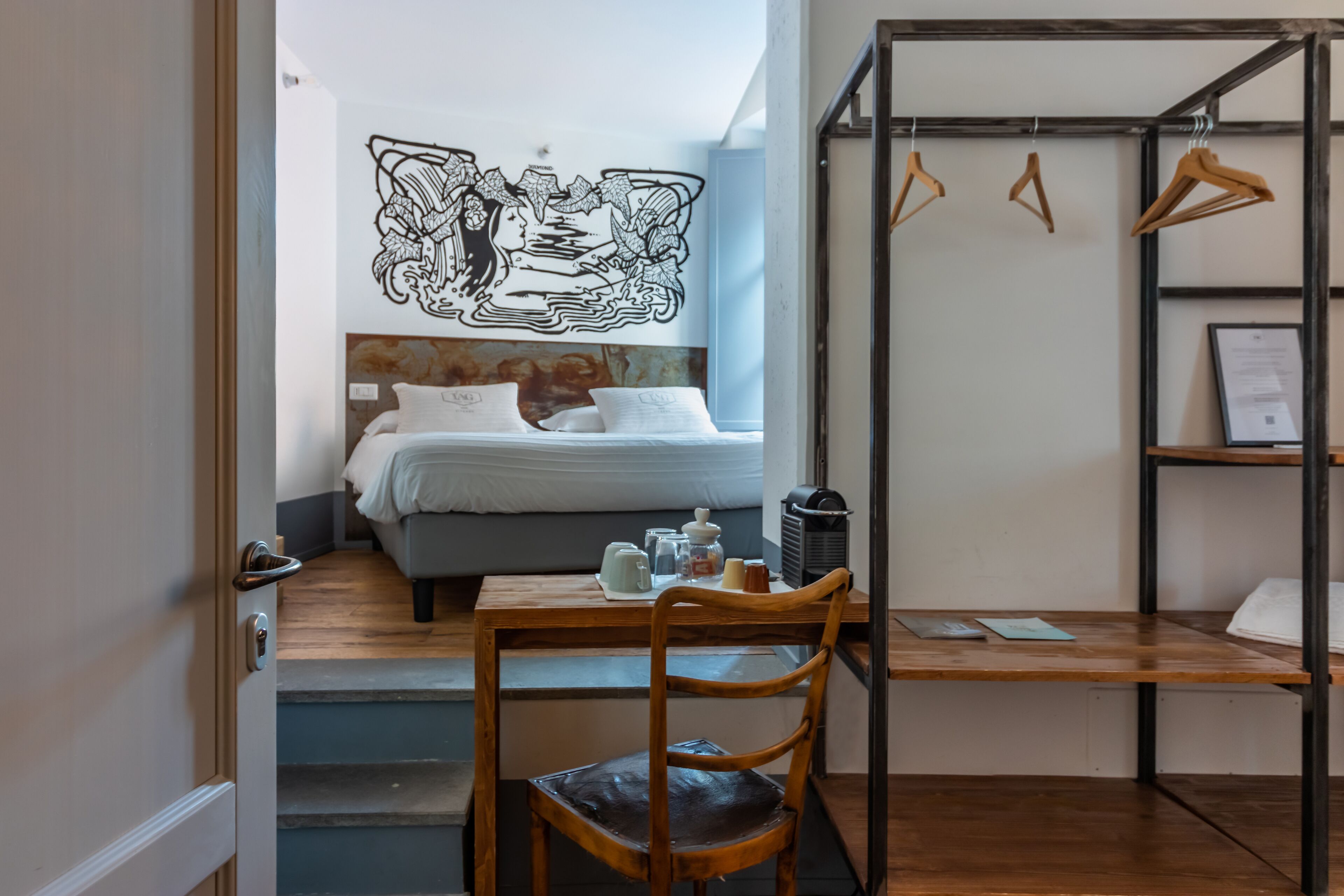 A compact hotel room with a bed, a wooden desk, and a chair. There is a coat rack with hangers and a framed certificate on the wall.