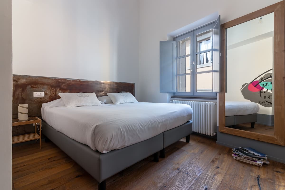 A bedroom with a wooden headboard, a large window with blue shutters, a radiator, and a mirror on the wall.