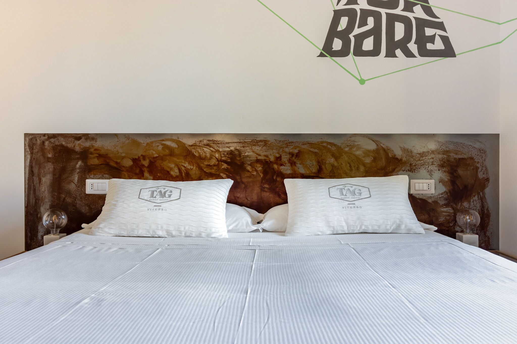 Un lit bien fait, avec une literie et des oreillers blancs, une tête de lit au motif texturé et un mur orné d’un motif géométrique et de l’inscription « TAC BARE ».