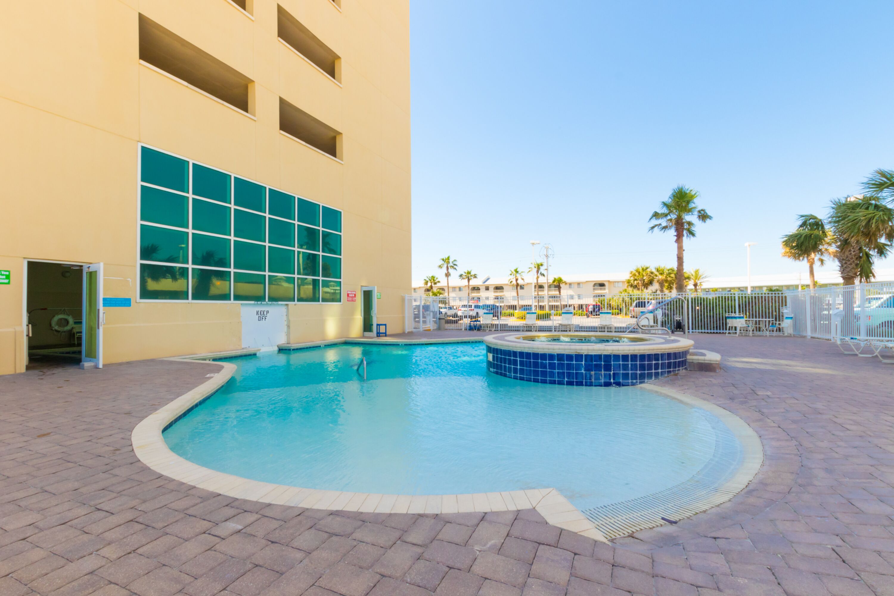 A swimming pool area with a hot tub, a covered seating area, and a building with large windows and a door labeled "KEEP OFF".