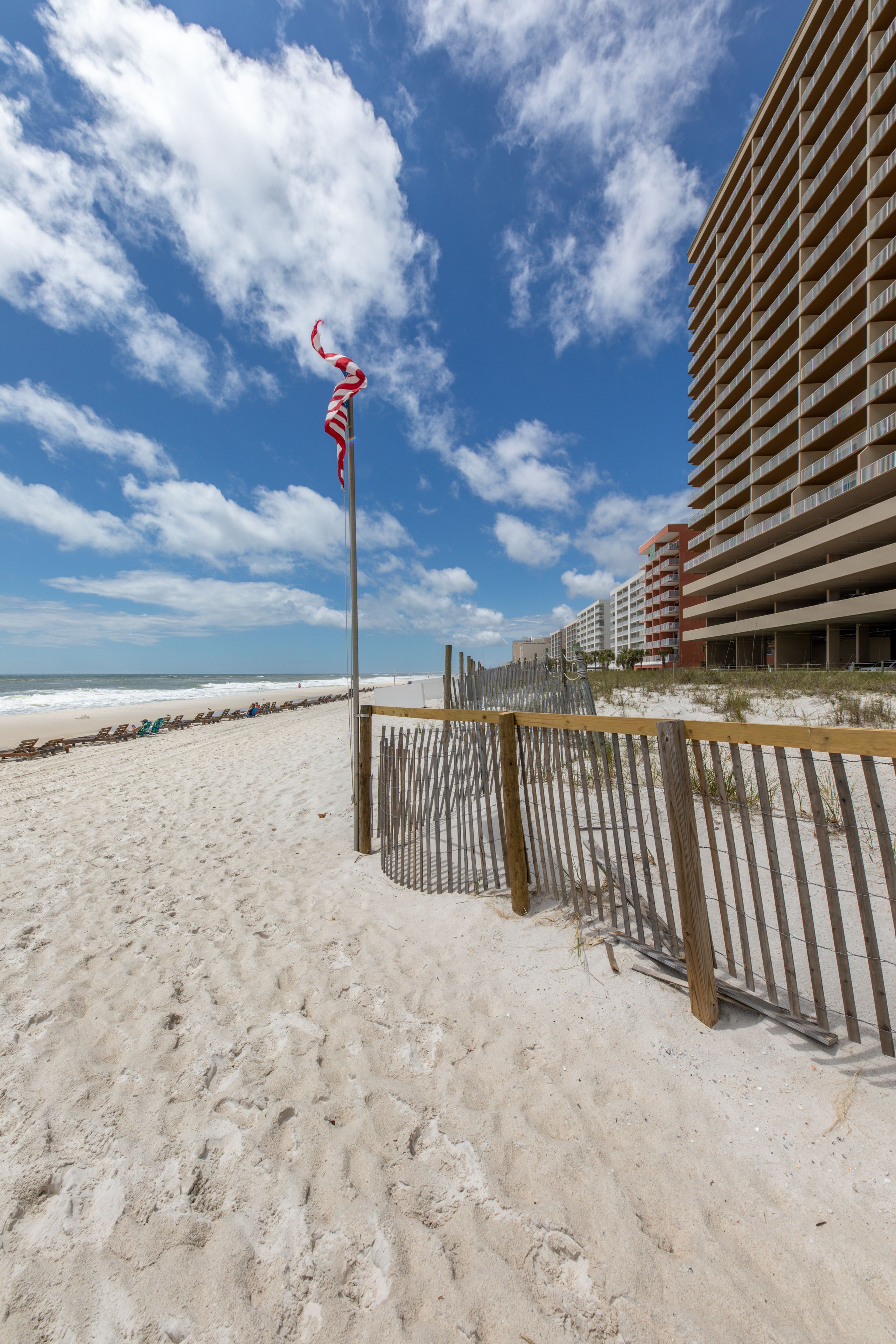 Une plage avec une clôture en bois, un mât avec un drapeau, et un hôtel en arrière-plan.