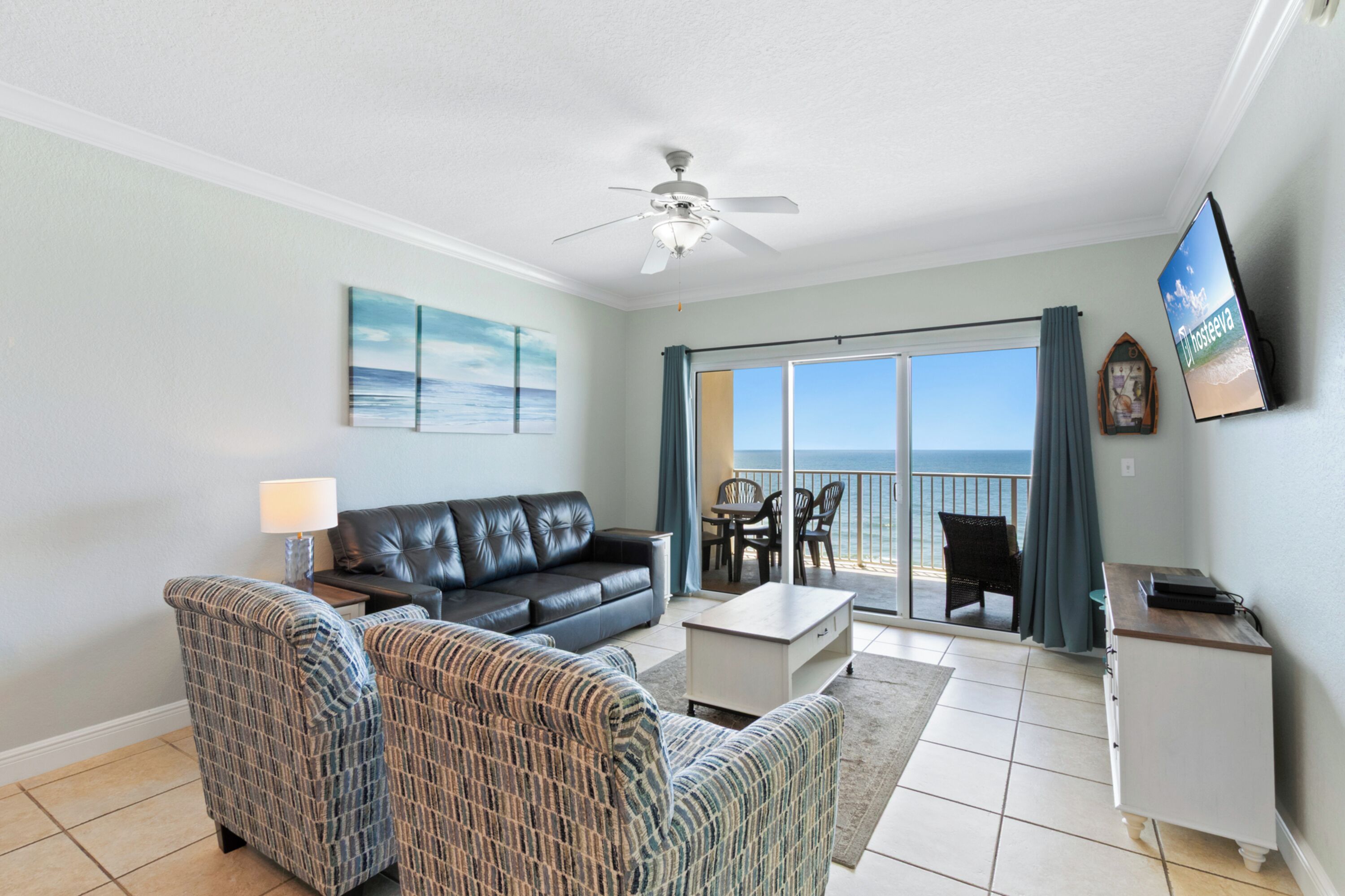 A living room with a black leather sofa, two patterned armchairs, a coffee table, and a TV on a stand. There is a ceiling fan, a painting on the wall, and a view of the ocean through a sliding glass door.