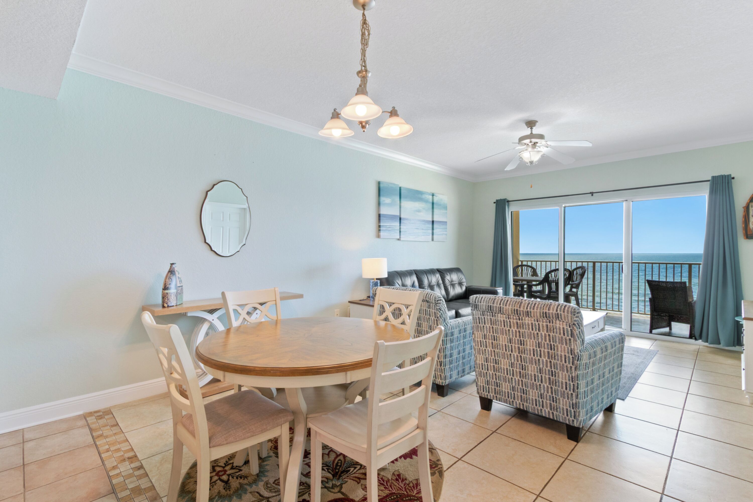 A dining area with a round table and chairs, a ceiling fan, and a view of the ocean through large windows.