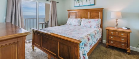 Un dormitorio con cama de madera, una mesita de noche, ventilador de techo y vistas al océano a través de un ventanal grande.