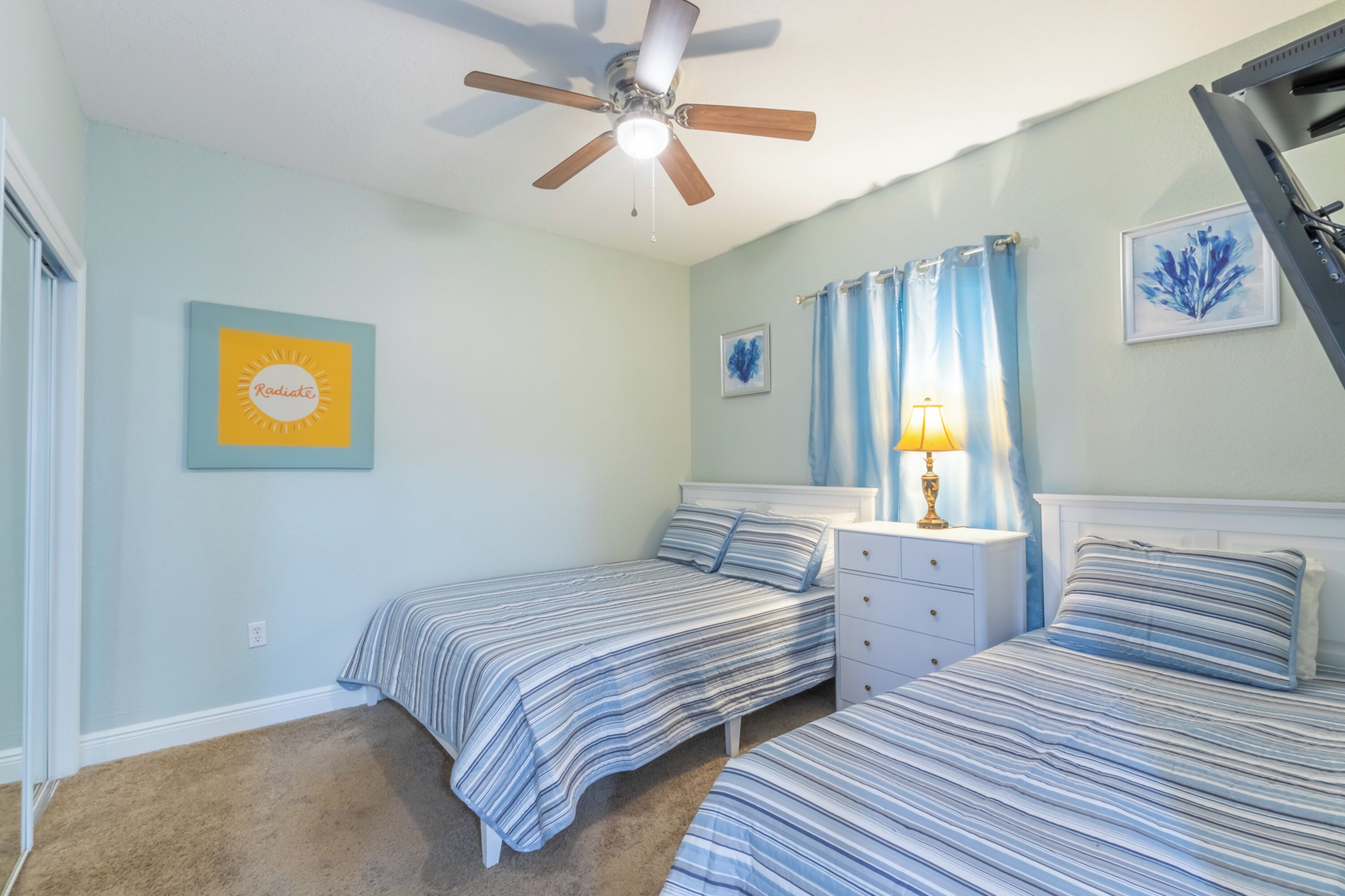 A bedroom with two beds, a ceiling fan, a nightstand with a lamp, and artwork on the walls.