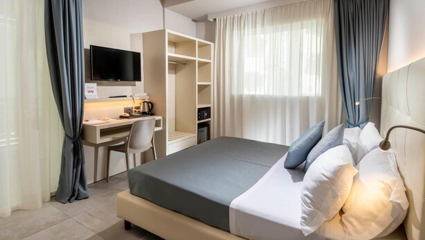 A modern hotel room with a bed, a desk with a computer, a television, and a wardrobe. - Hotel Manù (Rimini)