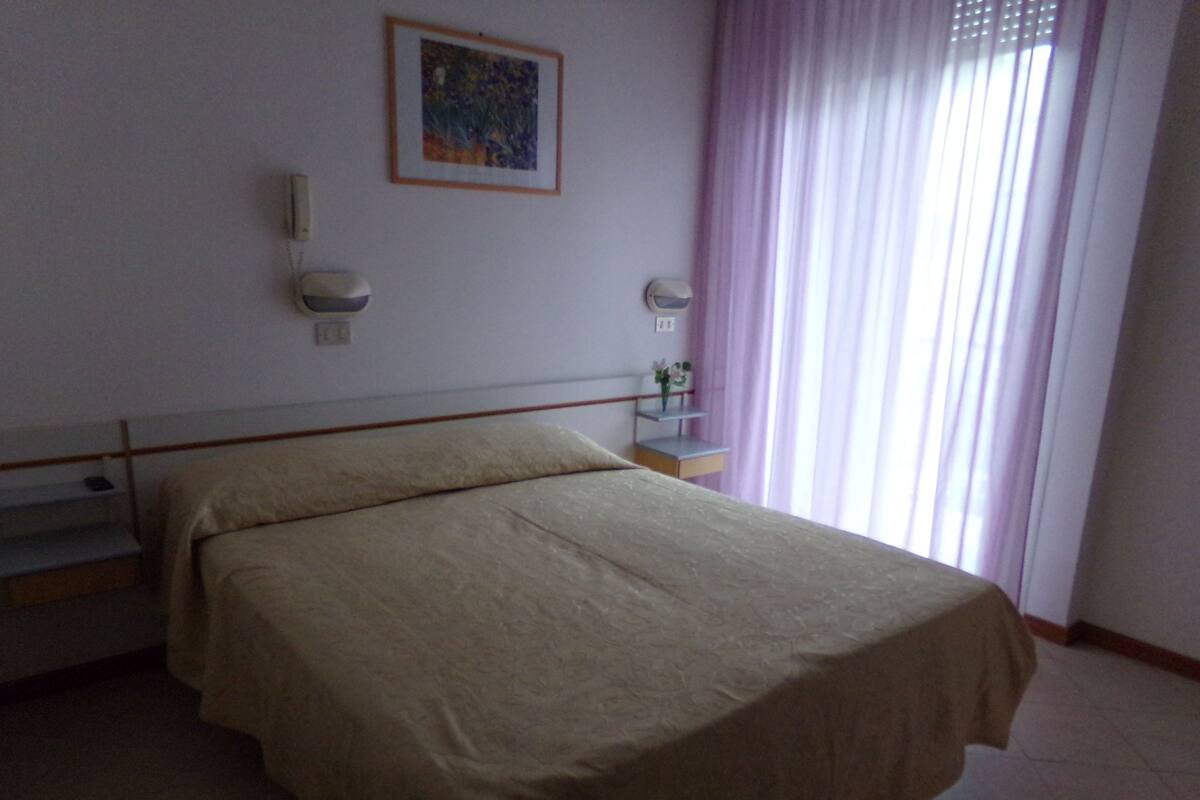 A bedroom with a bed, a nightstand, a wall-mounted lamp, and a window with curtains.