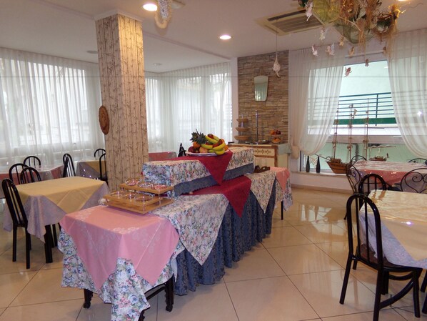 A dining area with tables set for a meal, a fruit display, and a view of the sea through large windows. - Hotel Manù (Rimini)
