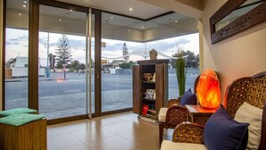 A modern hotel lobby with a glass door leading outside, a wooden reception desk, a seating area with cushions, and a decorative Buddha statue. - Desert Sands Boutique B&B - Self-Catering (Swakopmund)