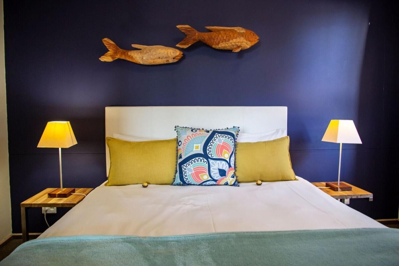 Ein Hotelzimmer mit einem großen Bett, zwei Nachttischlampen, einem farbenfrohen Kissen und zwei Fisch-Wandbildern.