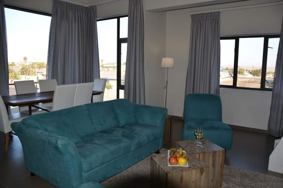 A modern living room with a teal sofa, a wooden coffee table with fruit, and a view of the city through large windows.