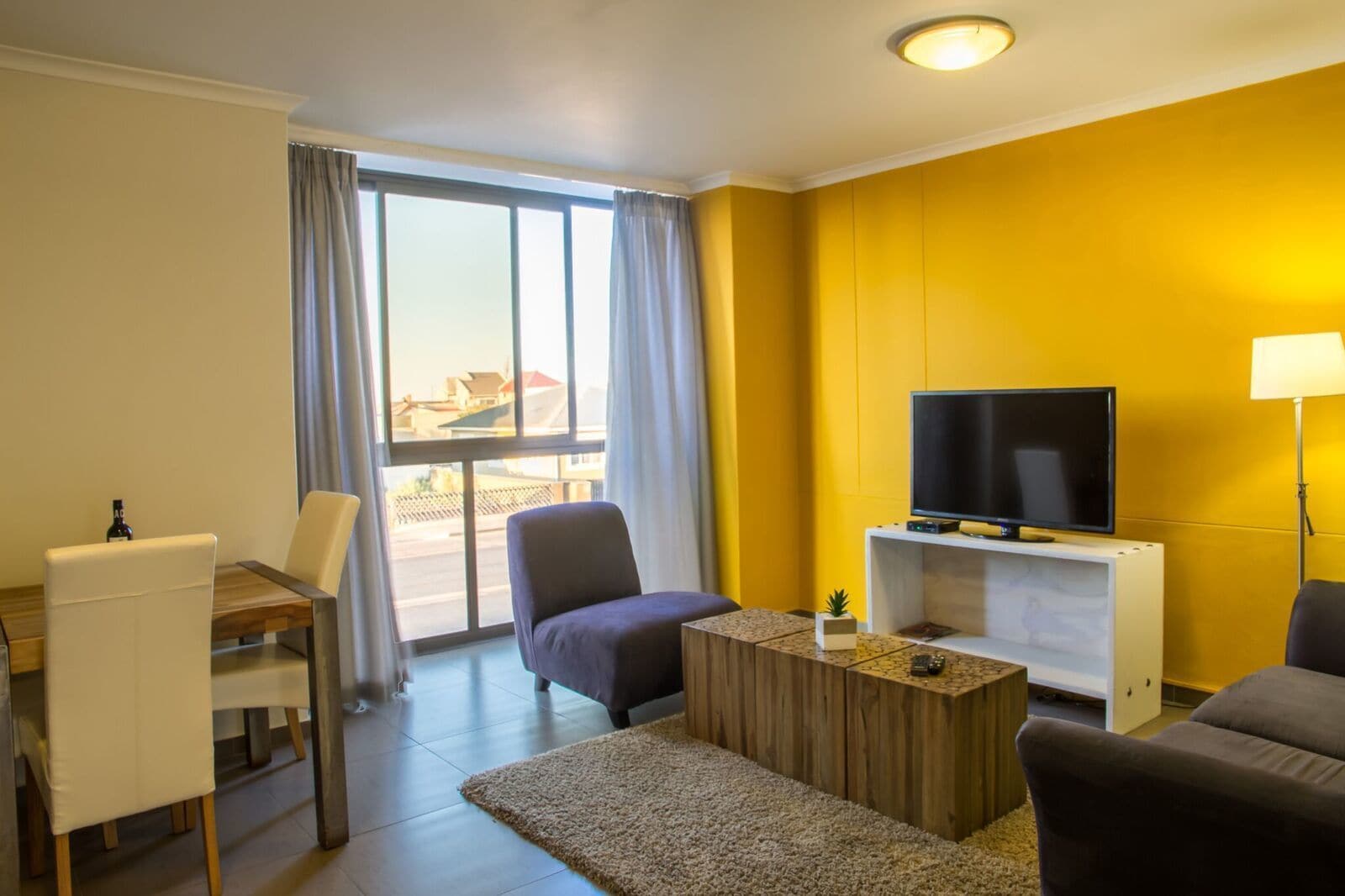 Habitación de hotel con televisión sobre un mueble, un sofá, una silla, una mesita y vistas al exterior a través de un ventanal grande.
