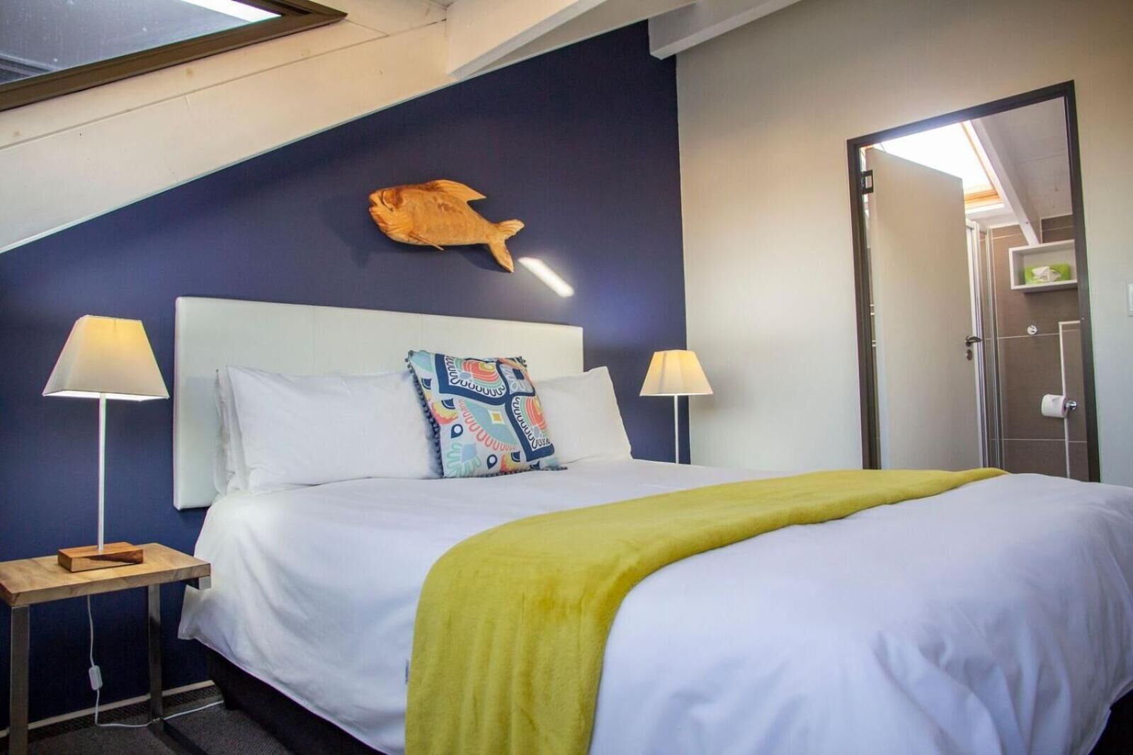A hotel room with a bed, two bedside tables, a nightstand, a colorful pillow, a wall-mounted fish decoration, and a door leading to a bathroom.