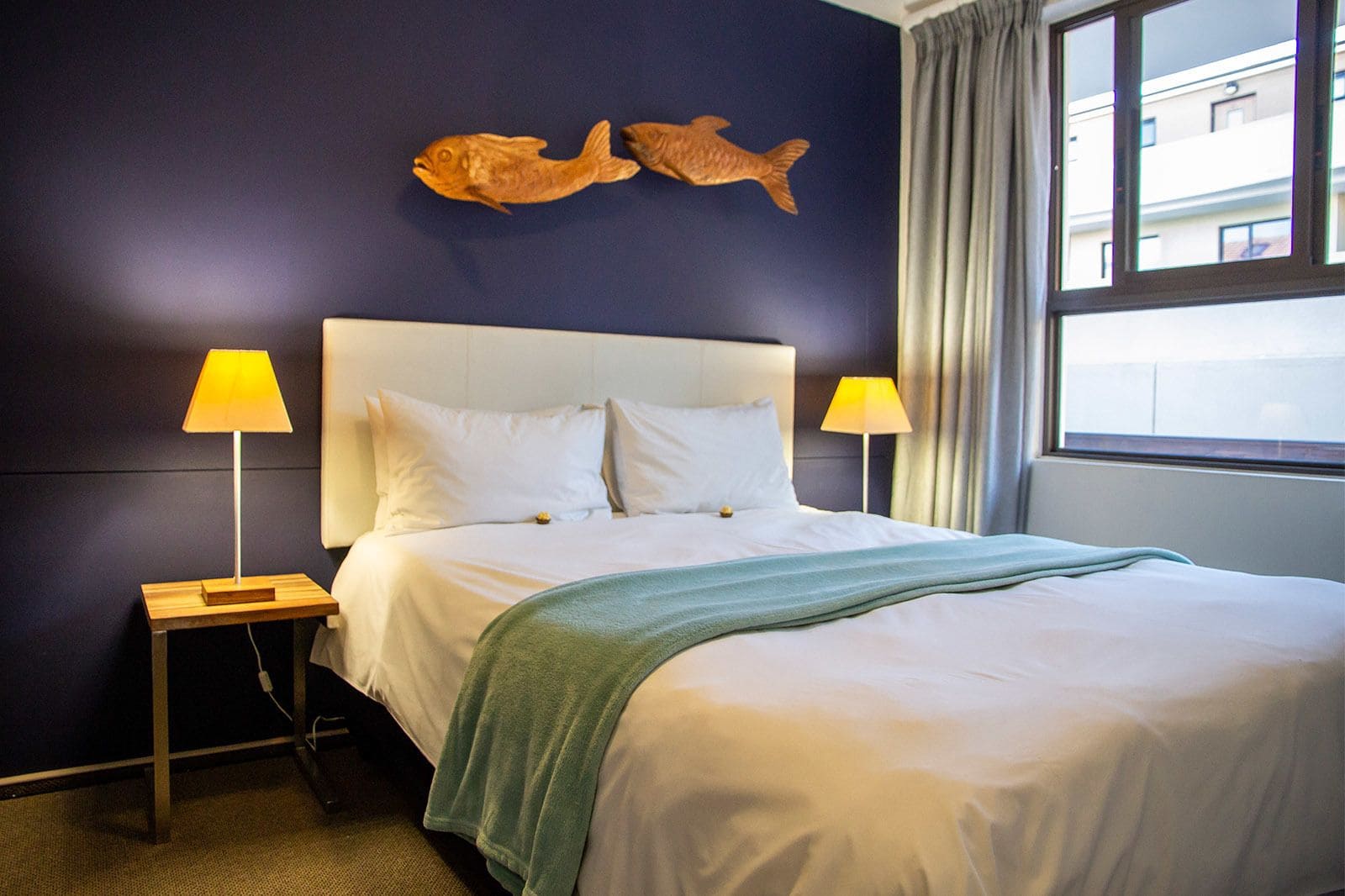 A hotel room with a bed, two bedside lamps, a green throw blanket, and two fish wall art pieces.