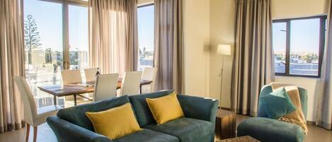 Ein modernes Hotelzimmer mit einem grünen Sofa, gelben Kissen, einem hölzernen Couchtisch und Blick auf die Stadt durch große Fenster.