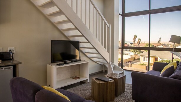 A modern living room with a TV on a white console, a staircase, and a view of a residential area through large windows. - Desert Sands Boutique B&B - Self-Catering (Swakopmund)