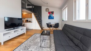 A modern living room with a flat-screen TV, a gray sofa, a dining area with a table and chairs, a staircase, and a kitchen area. - Lighthouse Apartments Tallinn (Tallinn)