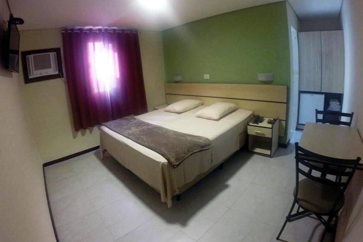 A bedroom with a bed, bedside table, chair, and a small kitchenette area.