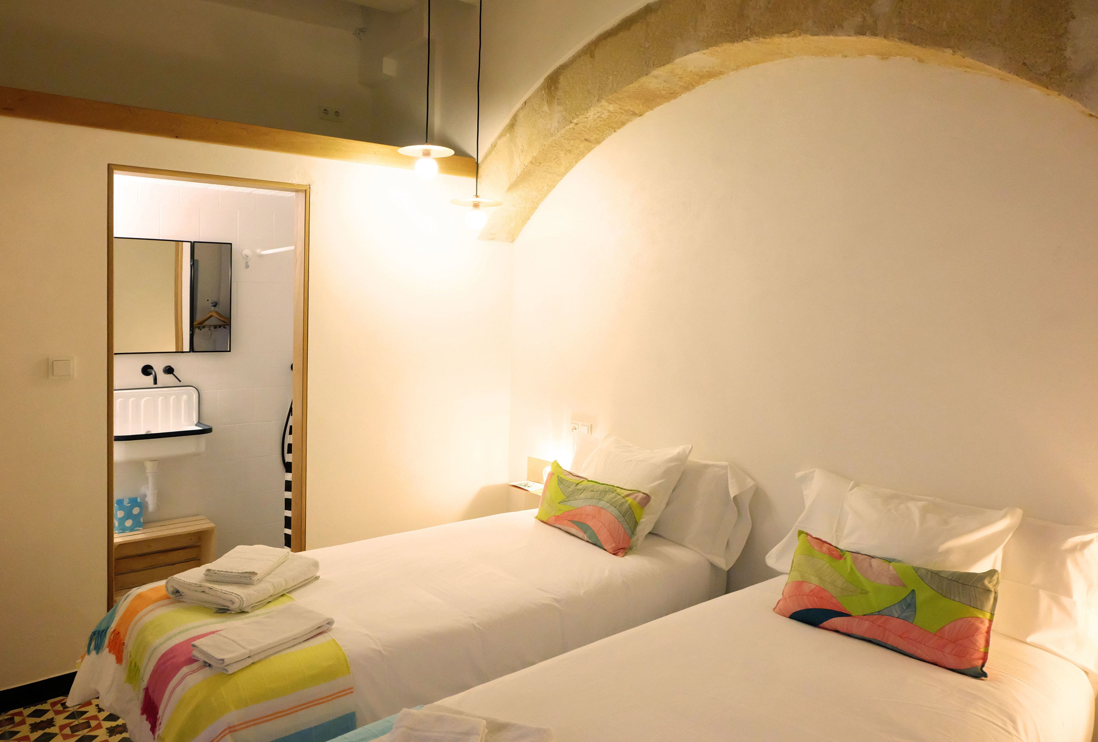 A hotel room with two beds, a mirror, a bedside table with a lamp, and a small bathroom area.