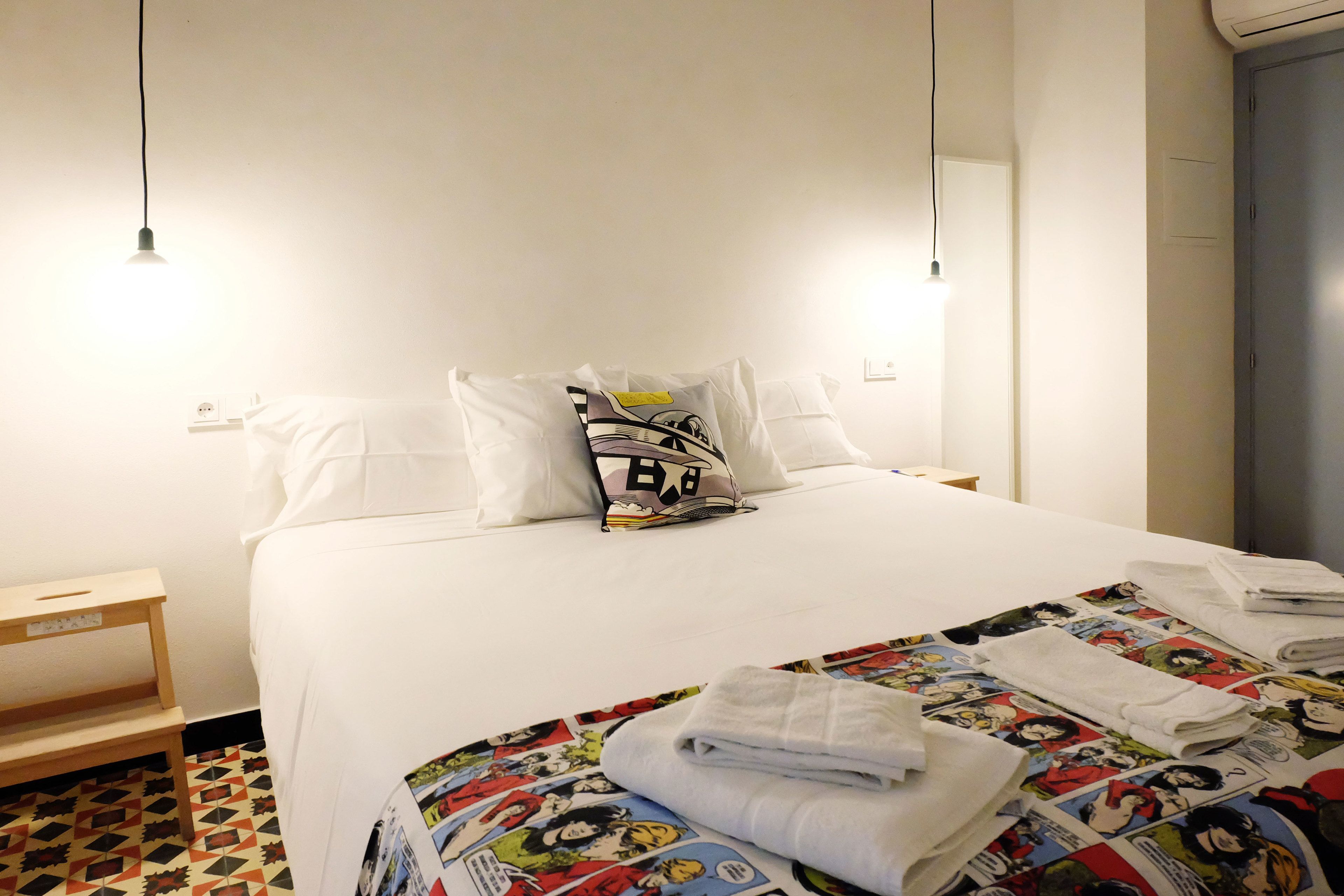 A hotel room with a neatly made bed, a small wooden nightstand, and a colorful comic book-themed bedspread.