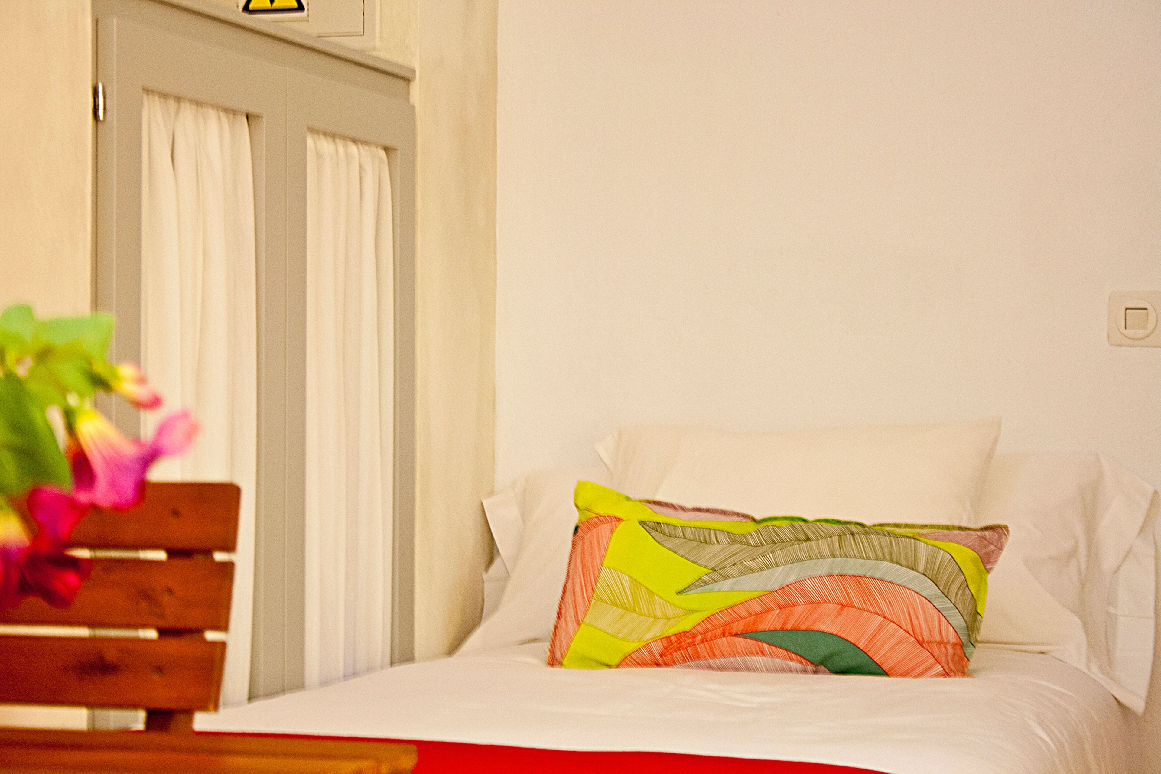 A neatly made bed with colorful pillows, a wooden bench, and a white door with curtains.