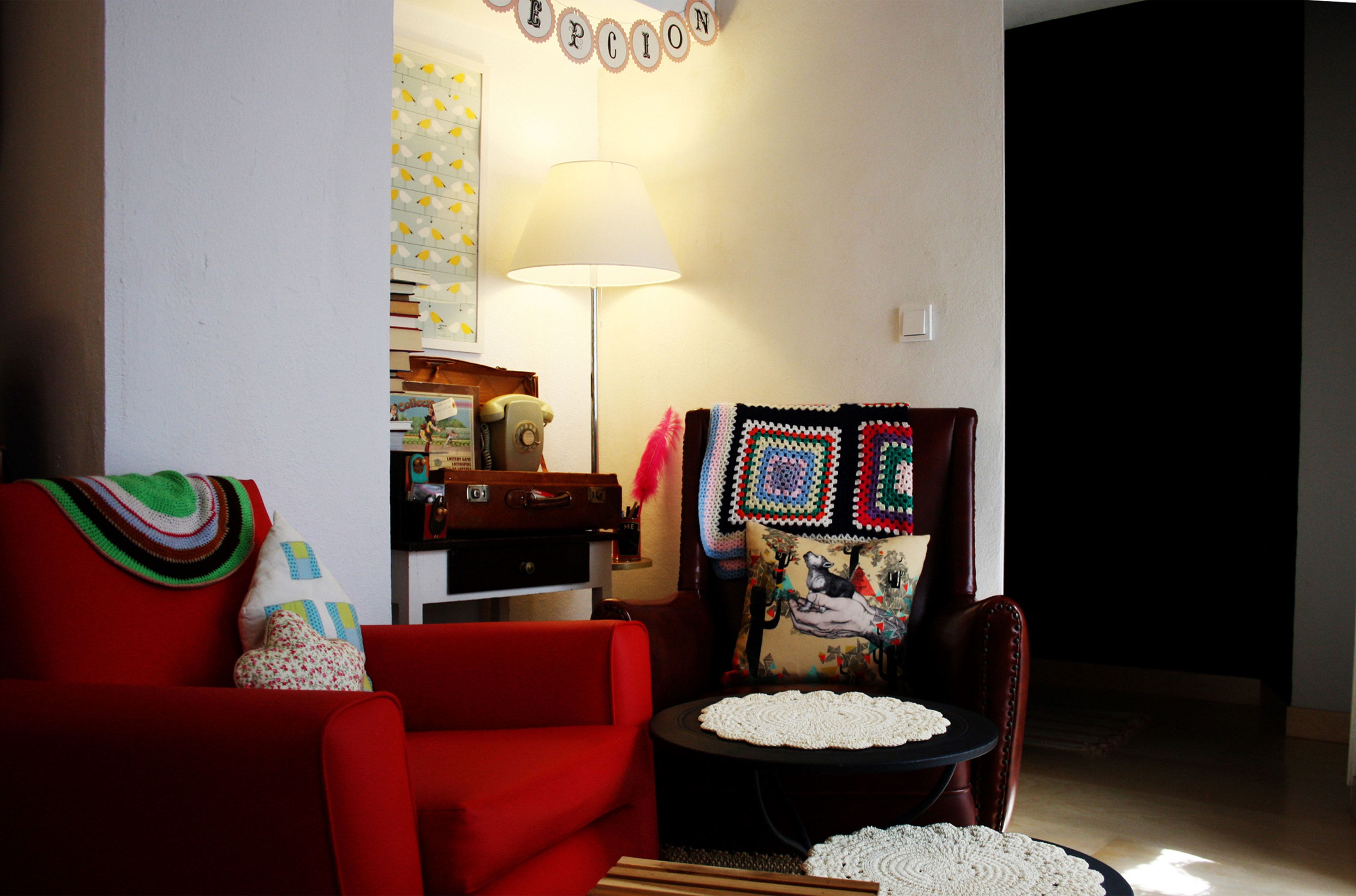 Una sala de estar con un sofá rojo, una mesita redonda con camino de mesa de ganchillo, una lámpara y un sillón con un cojín colorido.