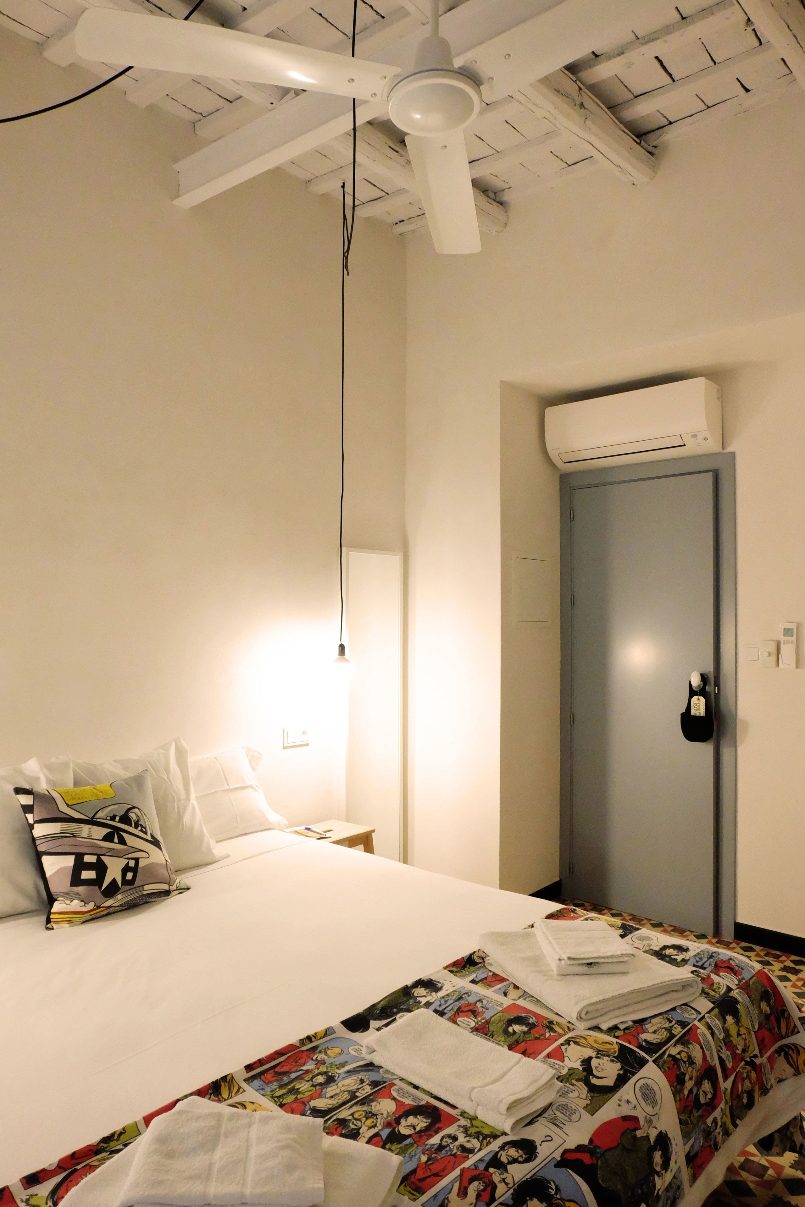 A hotel room with a bed, a ceiling fan, an air conditioning unit, and a door with a lock.