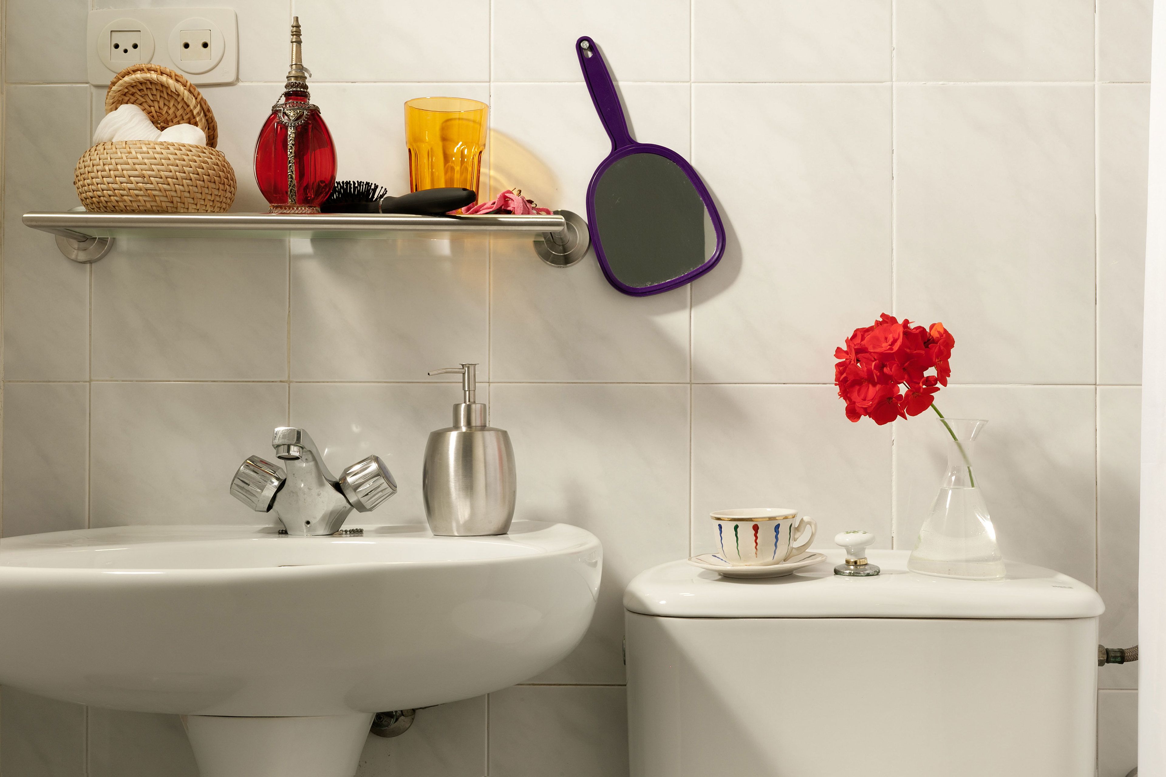 A bathroom with a white sink, a toilet, a shelf with a basket, a red vase, and a purple mirror.