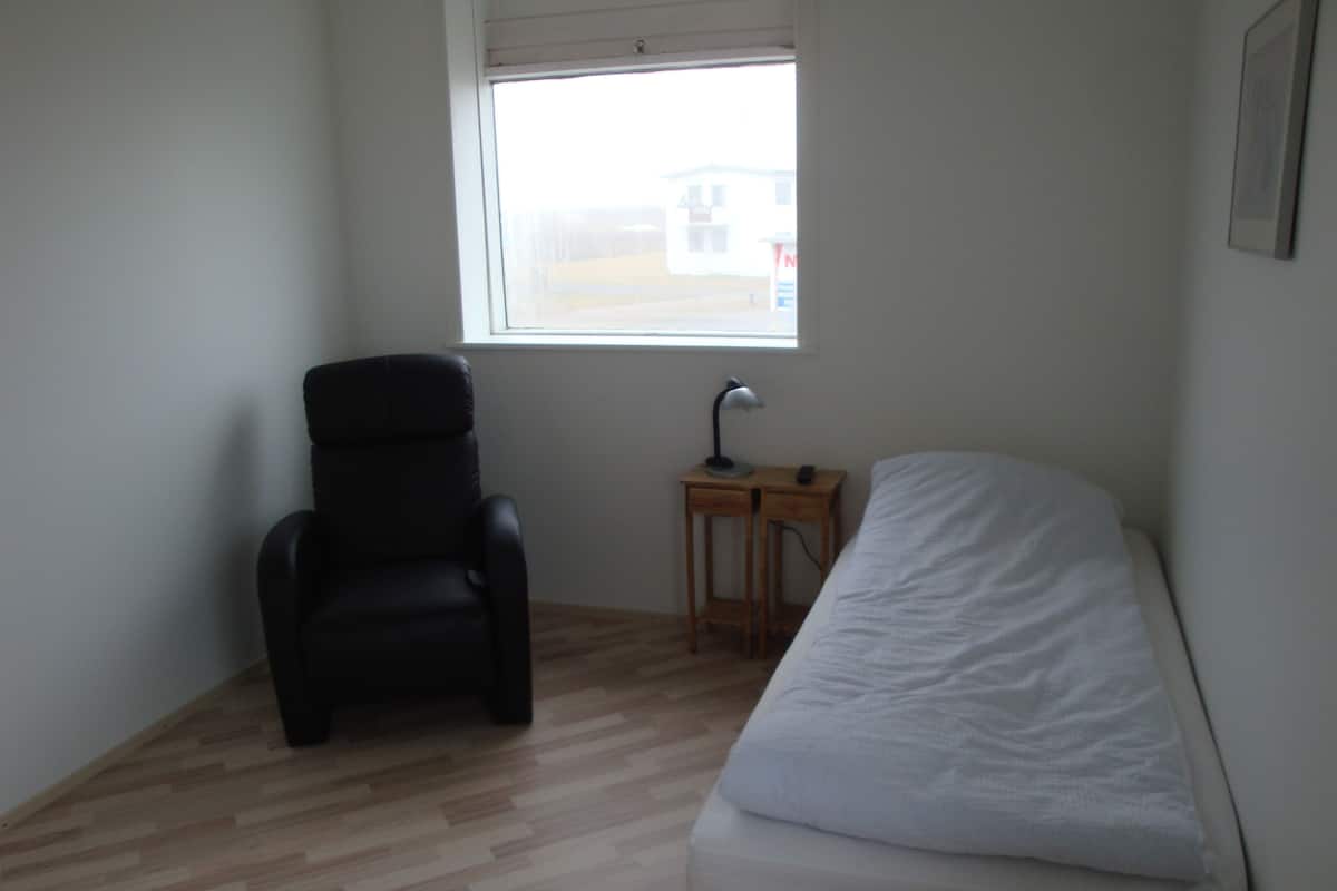 A small, sparsely furnished room with a single bed, a chair, a small table with a lamp, and a window with a view of a building outside.