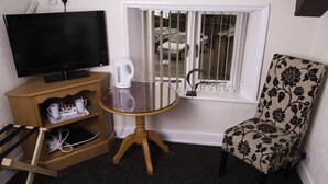 A room with a television on a wooden stand, a small round table with a white coffee pot, a floral-patterned chair, and a window with metal bars. - Fourways Guest House (Thirsk)
