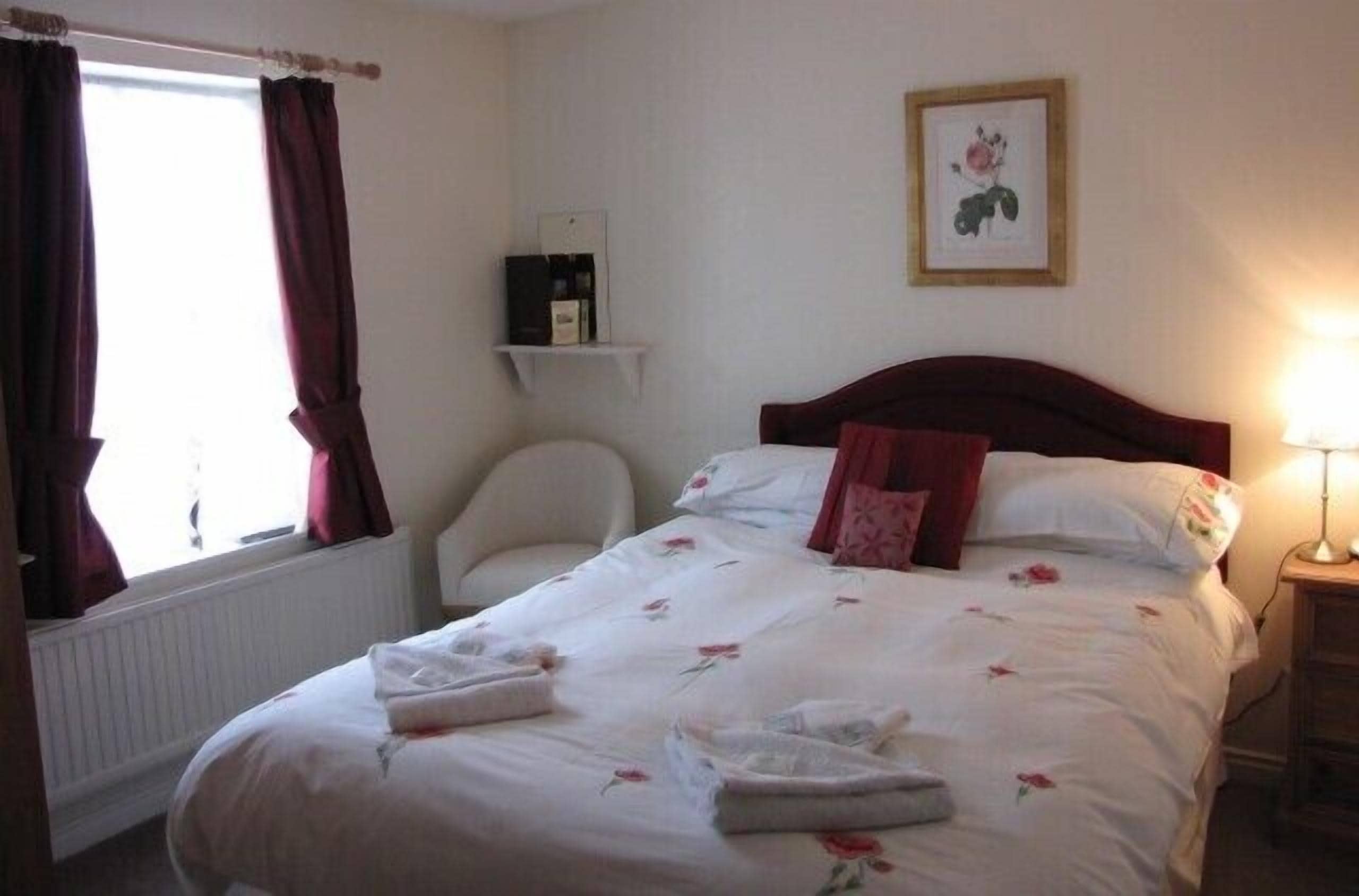 A hotel room with a bed, a chair, a bedside table with a lamp, a shelf with a framed picture, and a window with curtains.