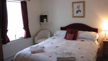 A hotel room with a bed, a chair, a bedside table with a lamp, a shelf with a framed picture, and a window with curtains.