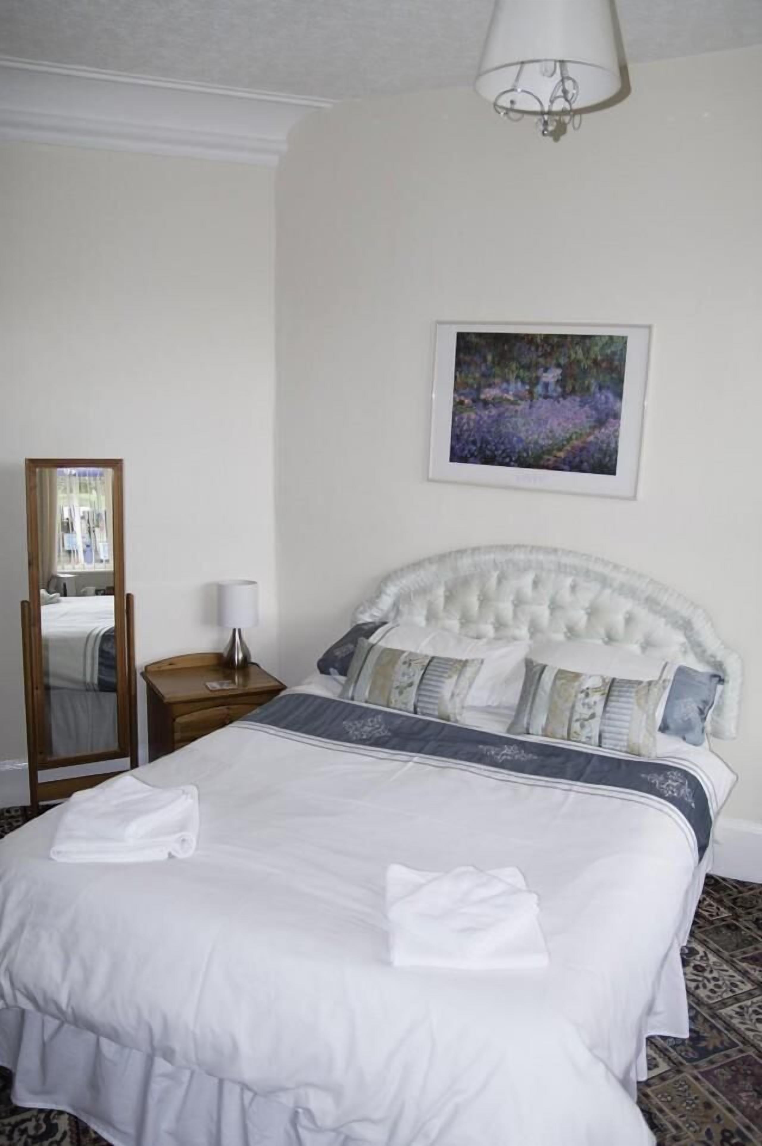 Un lit bien fait, avec du linge de lit blanc et une tête de lit, une table de chevet avec une lampe, un tableau encadré au mur et un miroir sur le mur.