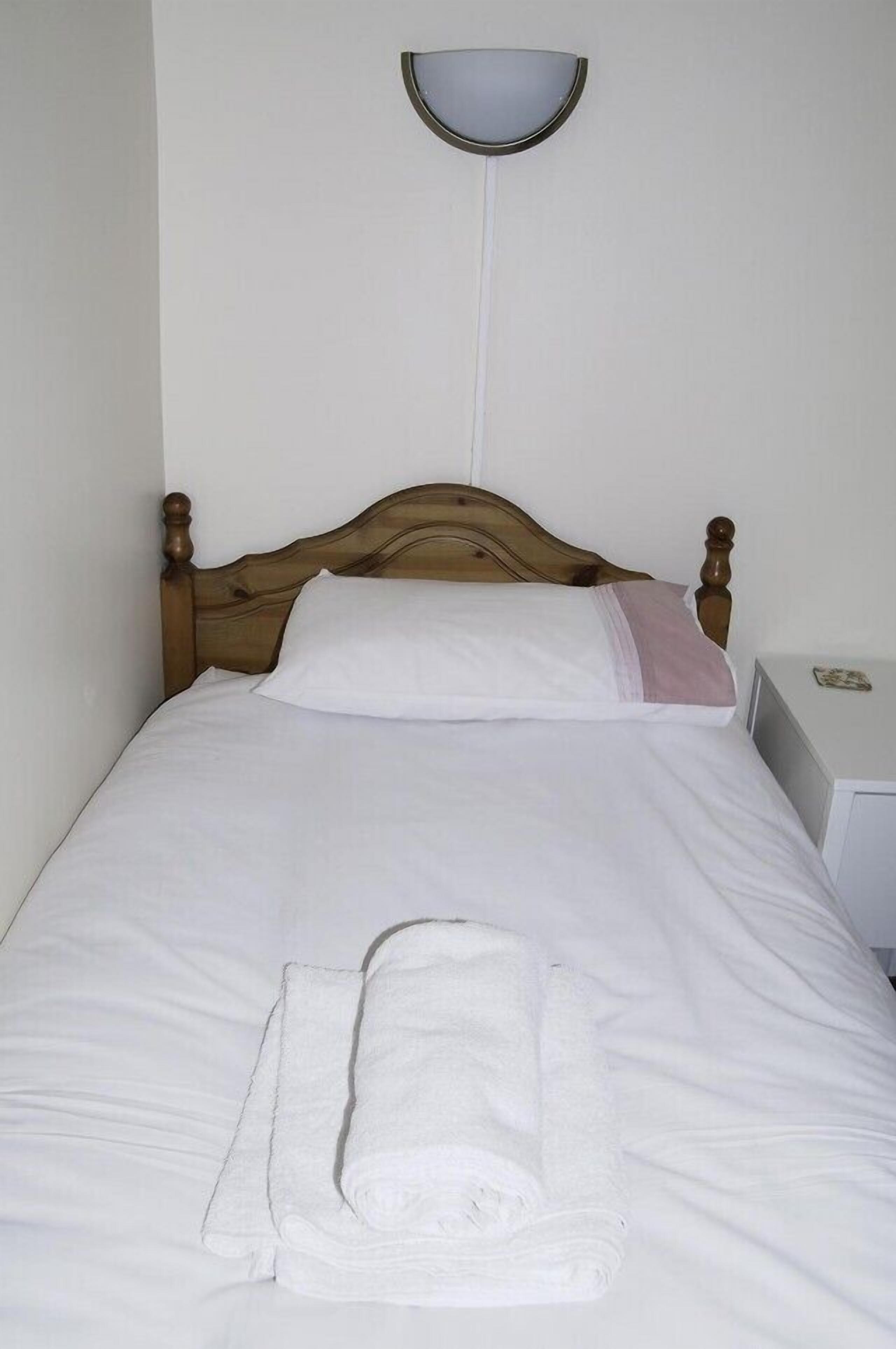 Una cama bien hecha con sábanas blancas y una toalla doblada, cabecera de madera, mesita de noche con lámpara y un aplique en la pared.