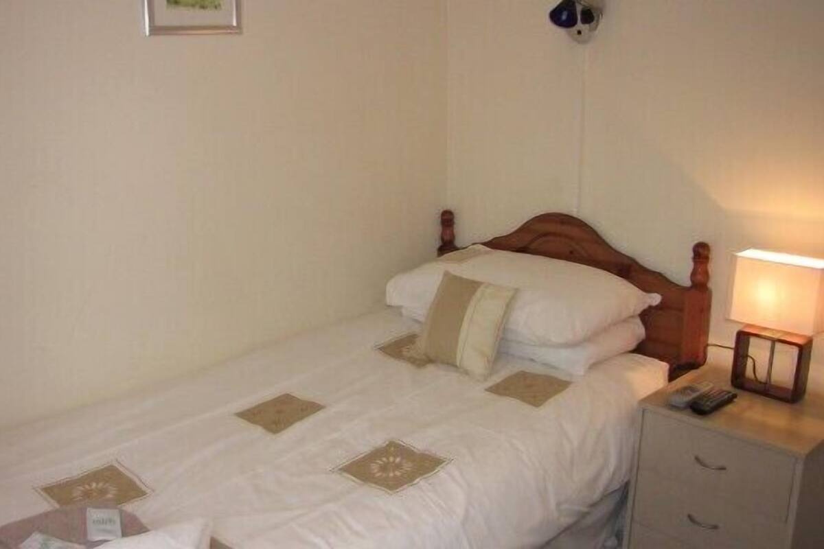 A single bed with a wooden headboard, a bedside table with a lamp, and a small drawer unit.