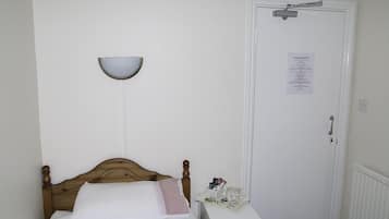 Una cama bien hecha con cabecera de madera, una mesita de noche con una taza y una bandeja de objetos, y una puerta blanca con un letrero.